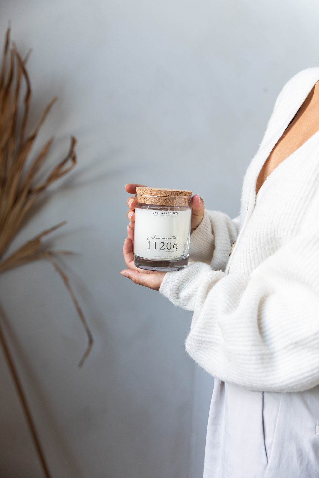 A person in a white ribbed cardigan holds a glass jar candle with a cork lid and a cream label reading '11206' against a soft gray wall with dried grass decor on the left.