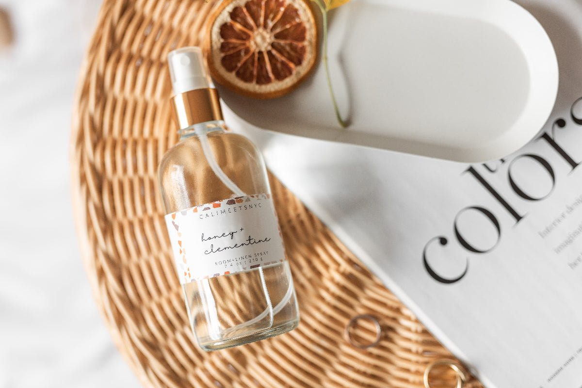 A clear glass spray bottle labeled 'Cali Meets NYC Honey + Clementine' with a gold collar and white spray top, styled on a woven wicker mat with a dried orange slice, a white ceramic dish, and a white magazine in the background.