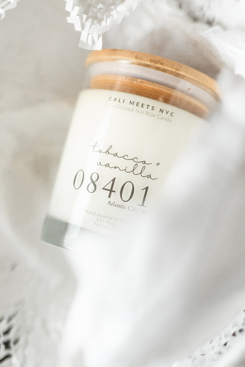 A close-up of a candle jar with a white wax body, a light wood lid, and a printed label reading 'CALI MEETS NYC' and 'tobacco + vanilla 08401'. The candle is photographed in soft white fabric packaging or wrapping.