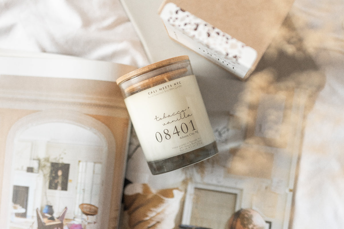A jar candle labeled 'tobscco vanilla 08401' by The Sweet Me, styled on top of a magazine page with a kraft gift box nearby.