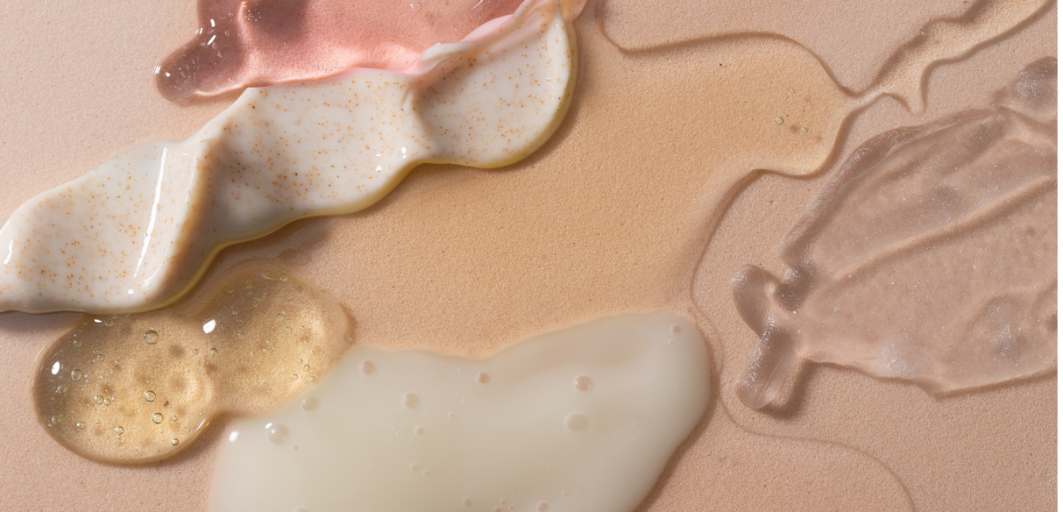 Close-up of several cosmetic creams and gels swirled on a beige surface, including a translucent pink cream, a thick white cream, a clear gel, and a golden-amber liquid.