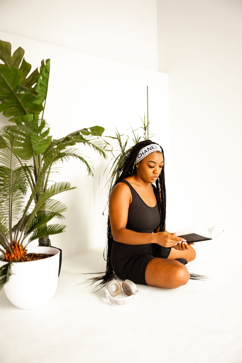 A woman with long black braids sits cross-legged on a white floor in a bright studio, wearing a black tank top and shorts, a white headband with black lettering, and white sneakers. She is holding a black tablet next to a large green potted plant.